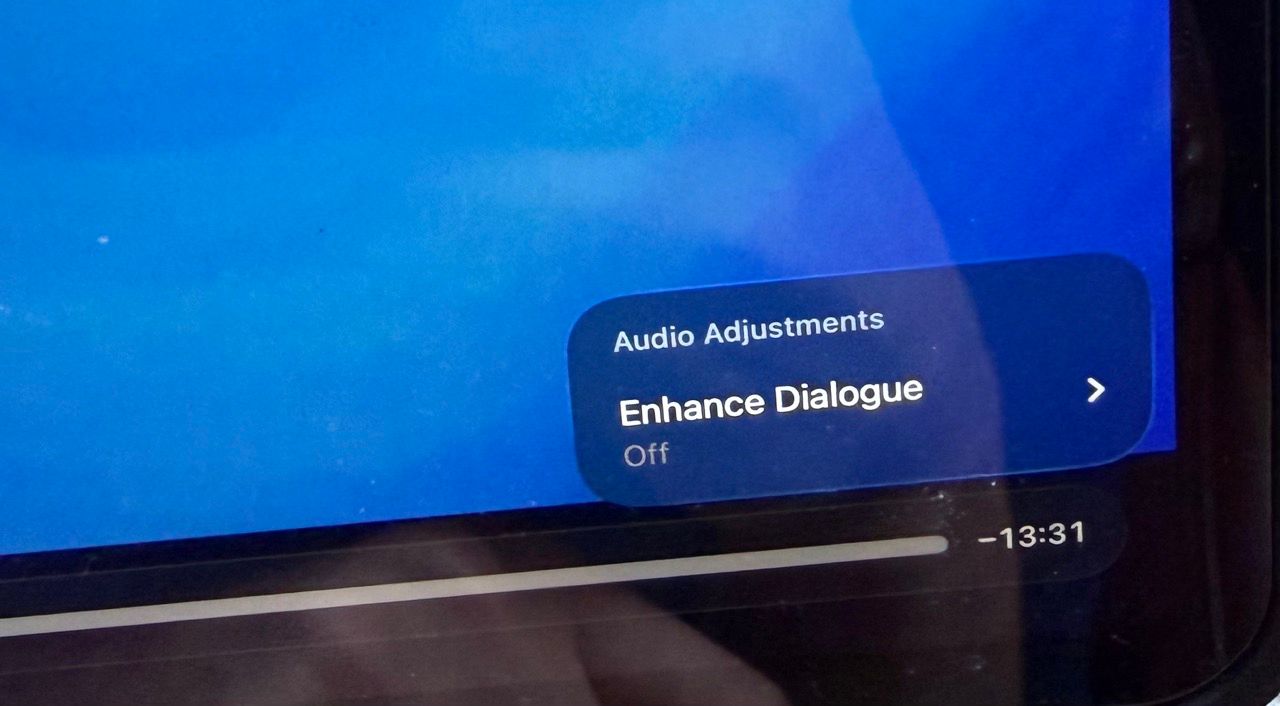A close-up shot of a digital screen displaying an Apple video interface menu titled Audio Adjustments. The specific setting Enhance Dialogue is visible and currently toggled to Off. A playback progress bar at the bottom shows 13 minutes and 31 seconds remaining in the video