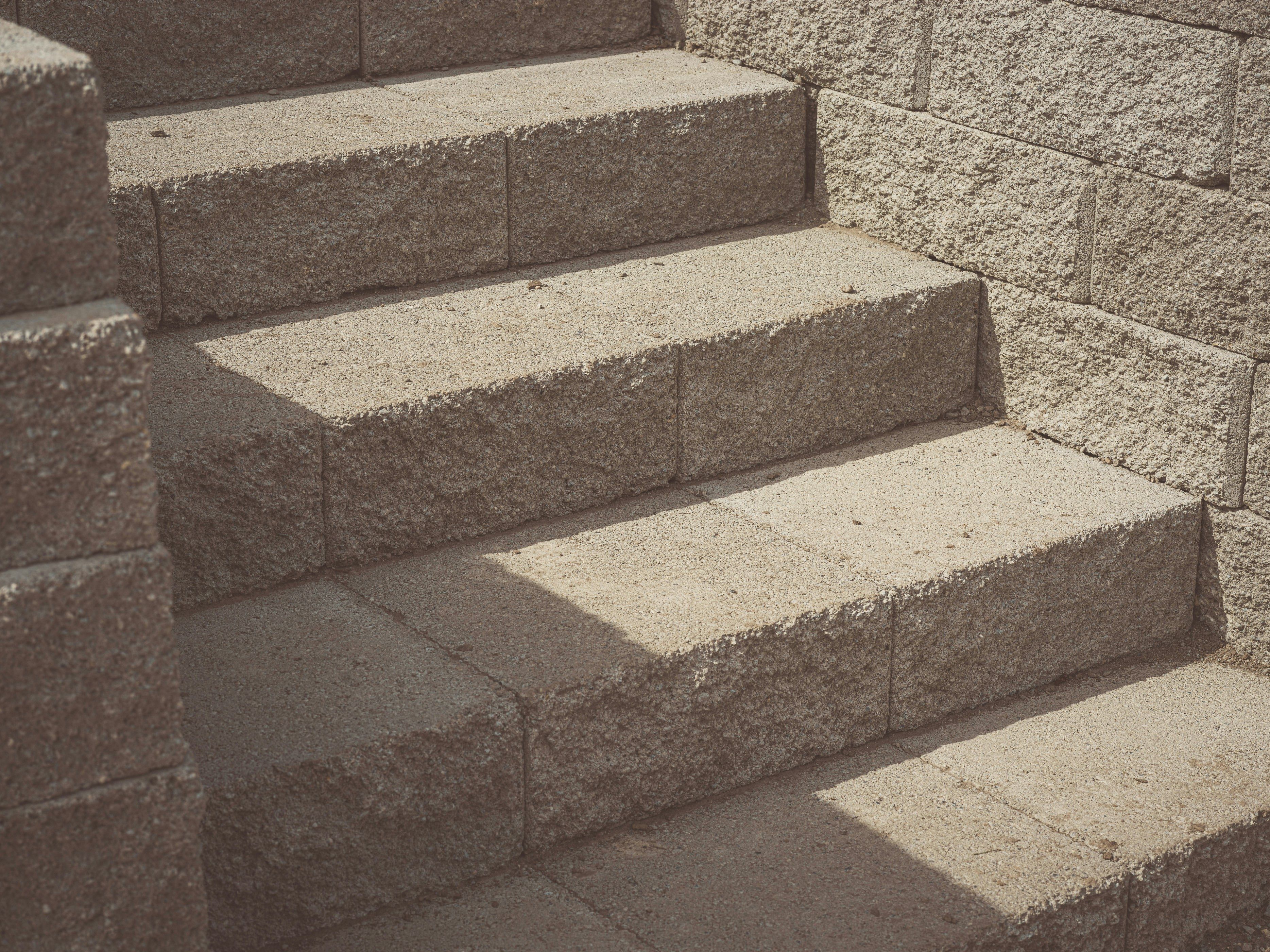 A low-angle, close-up shot of a set of stone steps made of rough-hewn, light-colored blocks. The steps represent different stages and ascend from the lower left to the upper right of the frame. Sunlight casts a sharp shadow diagonally across the lower two steps, highlighting the texture of the stone. The wall to the right is also constructed of the same type of stone blocks