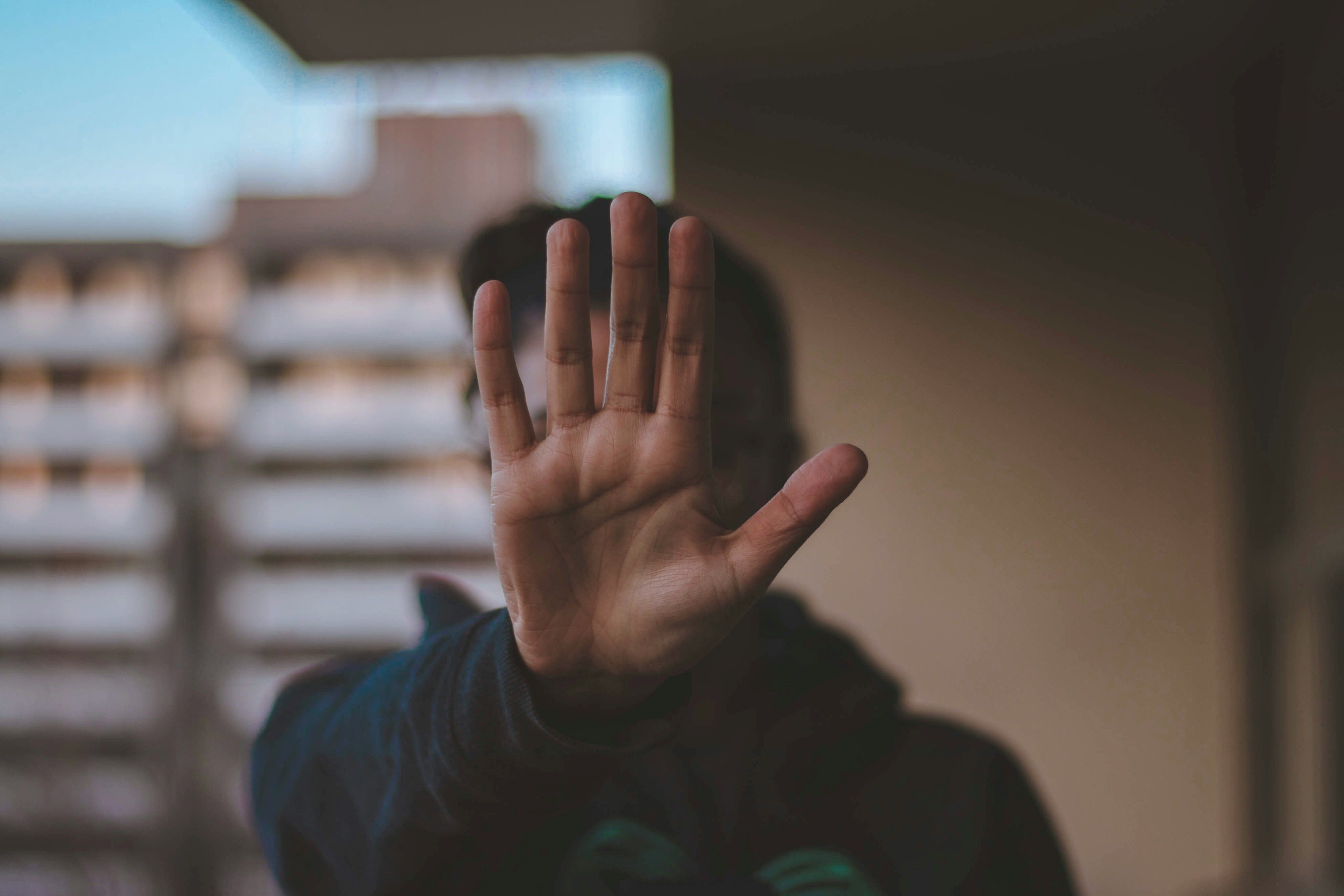 A person holds their hand up, palm facing the camera, in a gesture to stop. The background is a blurry building. The focus is on the hand, with the rest of the person and the background out of focus.