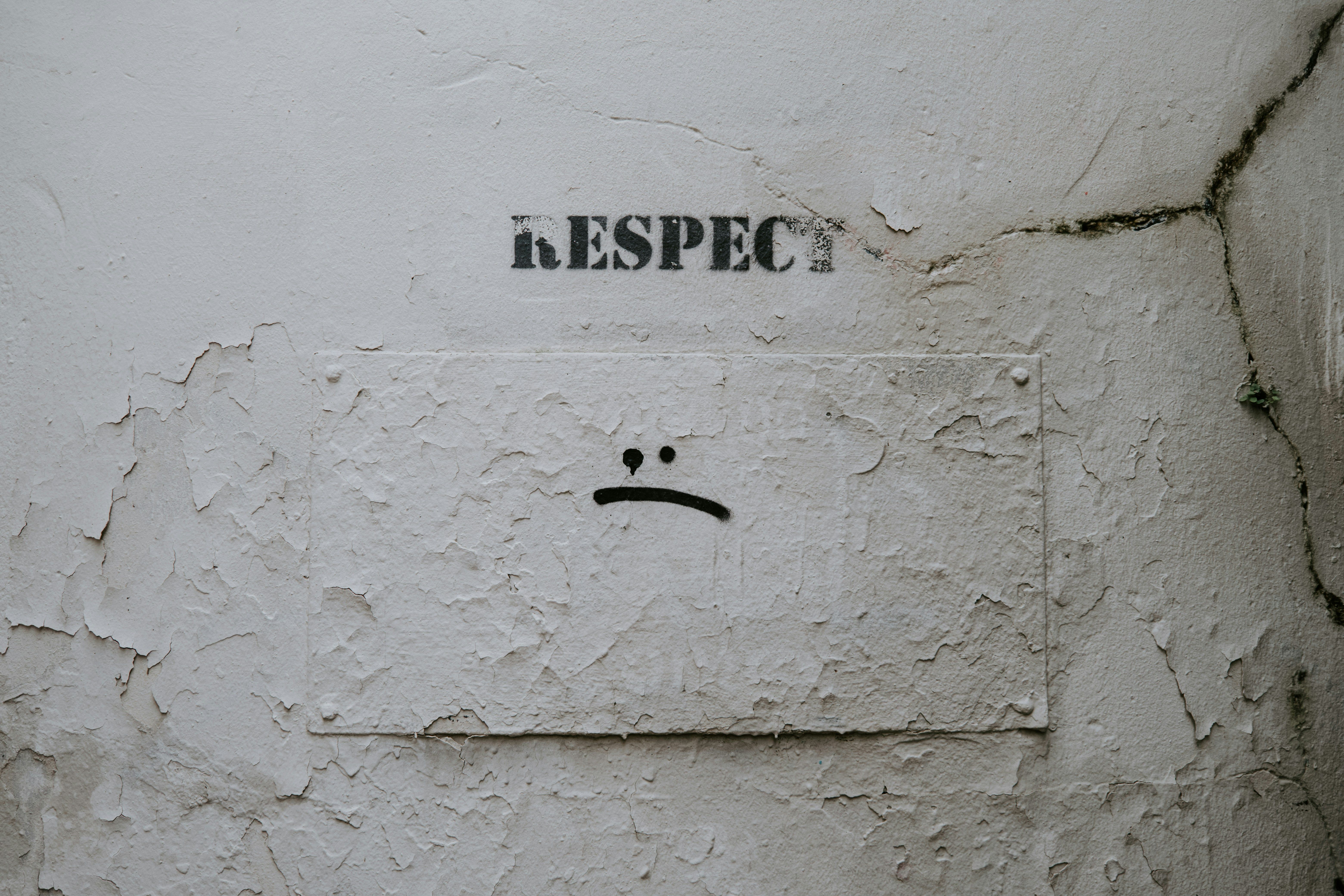 The text "RESPECT" is stencilled in black on a peeling, cracked white wall. Below it, a small, rectangular plaque is bolted to the wall, also in peeling white paint, with a black, stencilled frowny face.