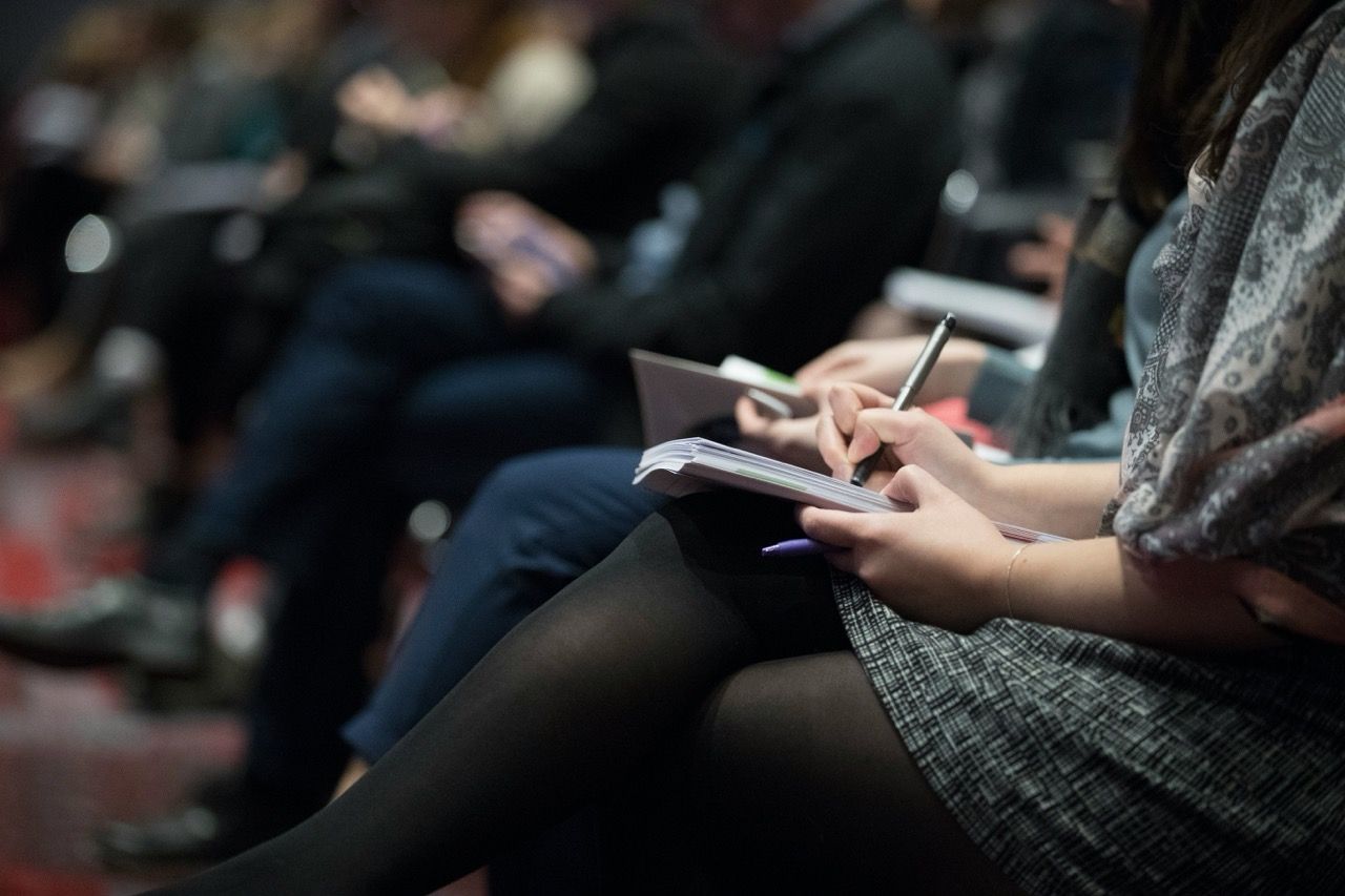 A close-up, shallow-depth-of-field shot of an audience member at a conference or lecture. A person in the foreground is seated with legs crossed, holding a notebook and a pen as if taking notes. The background is blurred, showing other seated attendees in a dimly lit auditorium setting