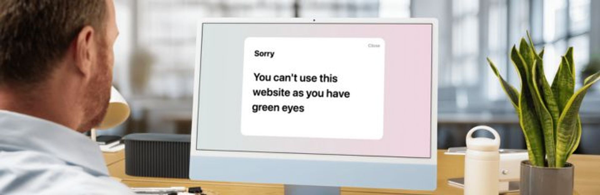 A white man with blondish hair in an office wearing a blue shirt, is looking at a light blue iMac visiting a website - the screen says: sorry you can't use this website as you have green eyes