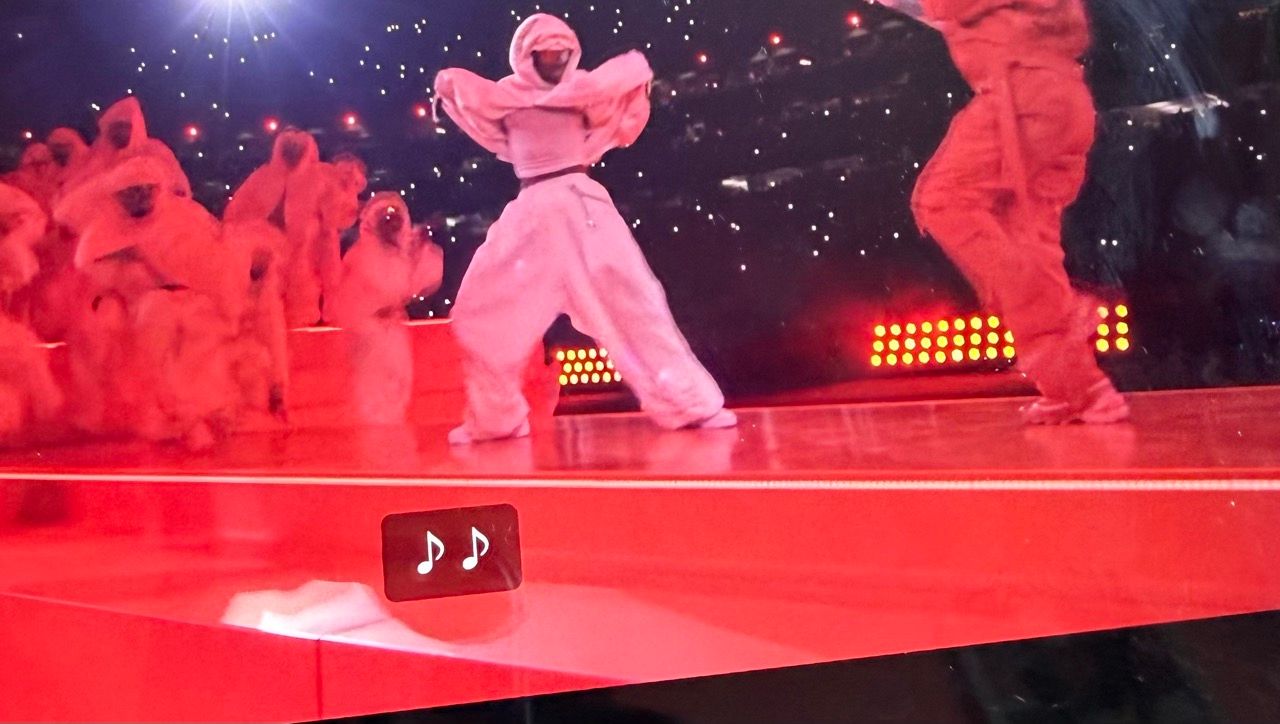 A wide shot of Rihanna performing at a past Super Bowl Halftime Show on a glowing red elevated platform. She is dressed in an all-red Loewe jumpsuit and puffer coat, surrounded by dancers in white hooded tracksuits. At the bottom center of the screen, a small black caption box is visible containing only two white musical note emojis (♪♪), illustrating the lack of descriptive or lyrical captions.