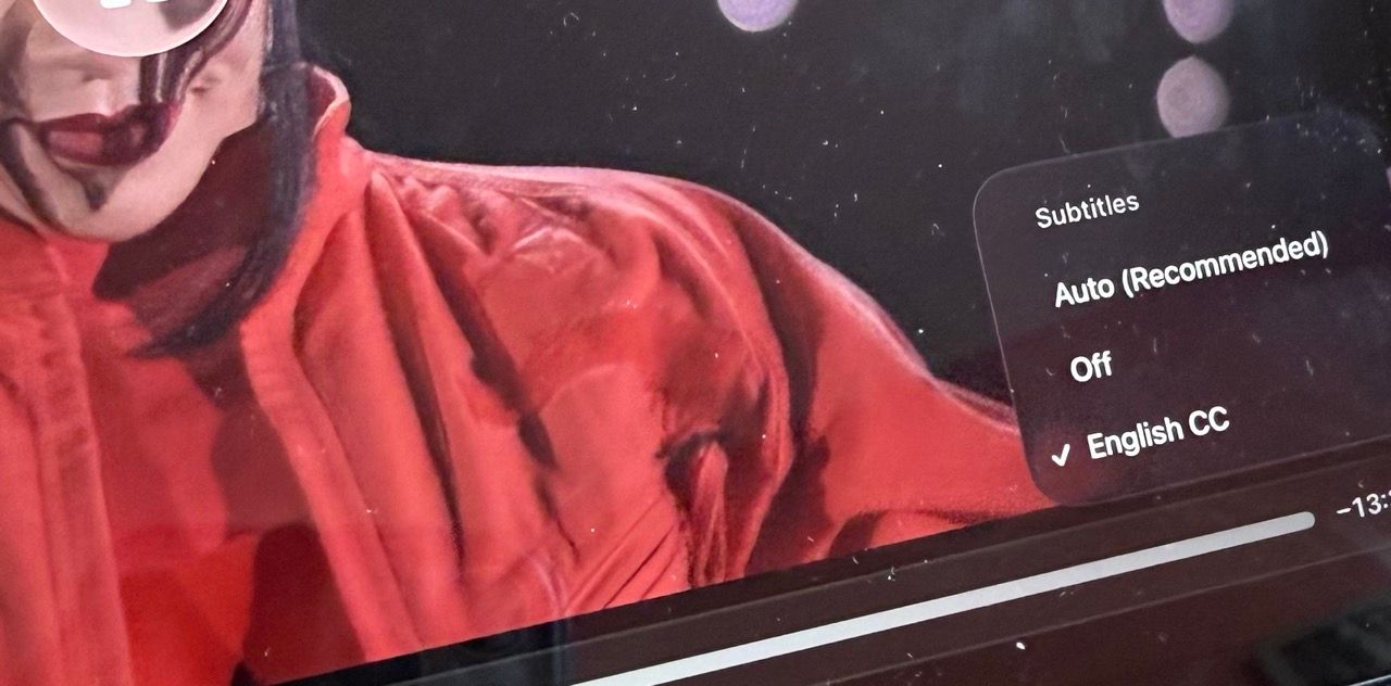 A close-up shot of a video player interface showing Rihanna in a bright red puffy Loewe jacket. On the right side of the screen, a Subtitles menu is visible, with English CC selected, indicated by a white checkmark. Other visible options in the menu include Auto (Recommended) and Off. A progress bar at the bottom indicates the video is currently playing.