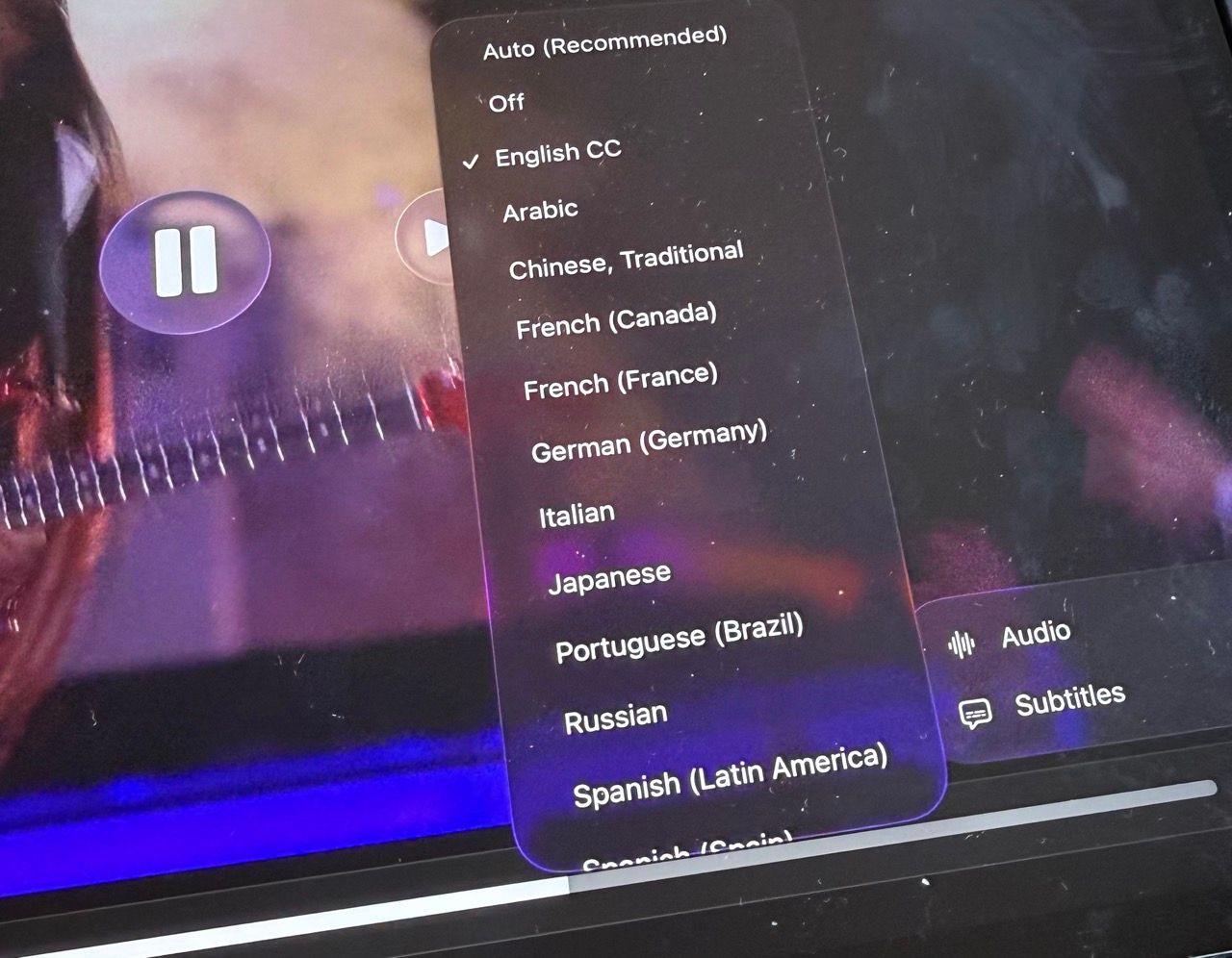 A close-up, angled photo of a tablet screen showing a video player's subtitle settings menu. A semi-transparent overlay lists various language options, with English CC currently selected with a checkmark. Other visible options include Arabic, Chinese, French, German, Italian, Japanese, Portuguese, Russian, and Spanish. In the background, a blurred video of a person playing a guitar is visible behind the menu.