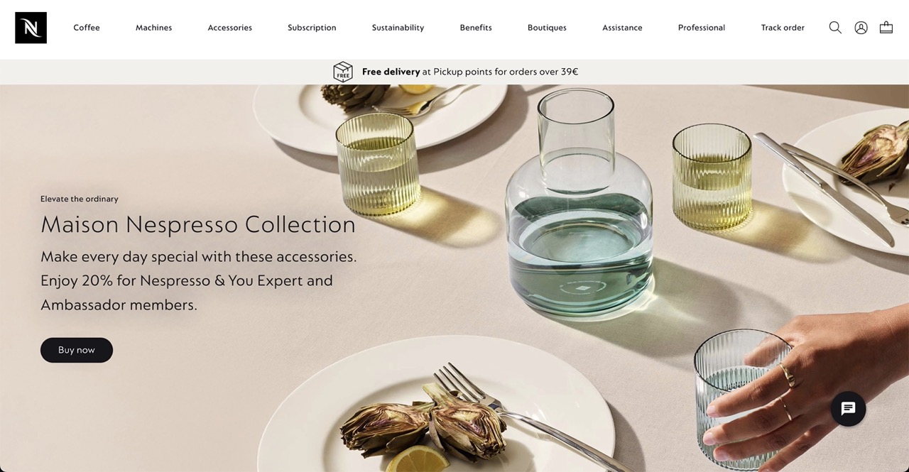 A Nespresso homepage for the Maison Nespresso Collection, featuring a sophisticated dining table scene with a clear glass carafe, ribbed tumblers, plates, silverware, and prepared artichokes with lemon slices. A hand reaches for one of the glasses in the foreground. Text overlay promotes the collection: Elevate the ordinary Maison Nespresso Collection. Make every day special with these accessories. Enjoy 20% for Nespresso & You Expert and Ambassador members, with a Buy now button. The top of the page includes the Nespresso logo and navigation menu, along with a free delivery offer.