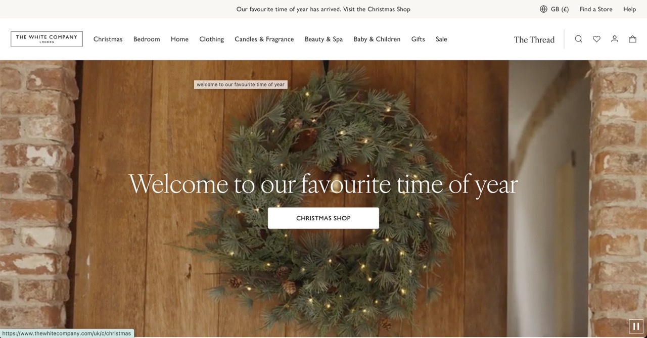 Screenshot of The White Company website promoting its Christmas collection, featuring a large, realistic Christmas wreath with small lights and pinecones hanging on a rustic wooden door, which is framed by brick and stone archways. The central message overlaid on the image reads Welcome to our favourite time of year, with a button directing users to the CHRISTMAS SHOP. Above the main image, the website's navigation is visible, including the brand's logo, links for categories like Christmas, Bedroom, Home, and Candles & Fragrance, and a top banner announcing, Our favourite time of year has arrived. Visit the Christmas Shop
